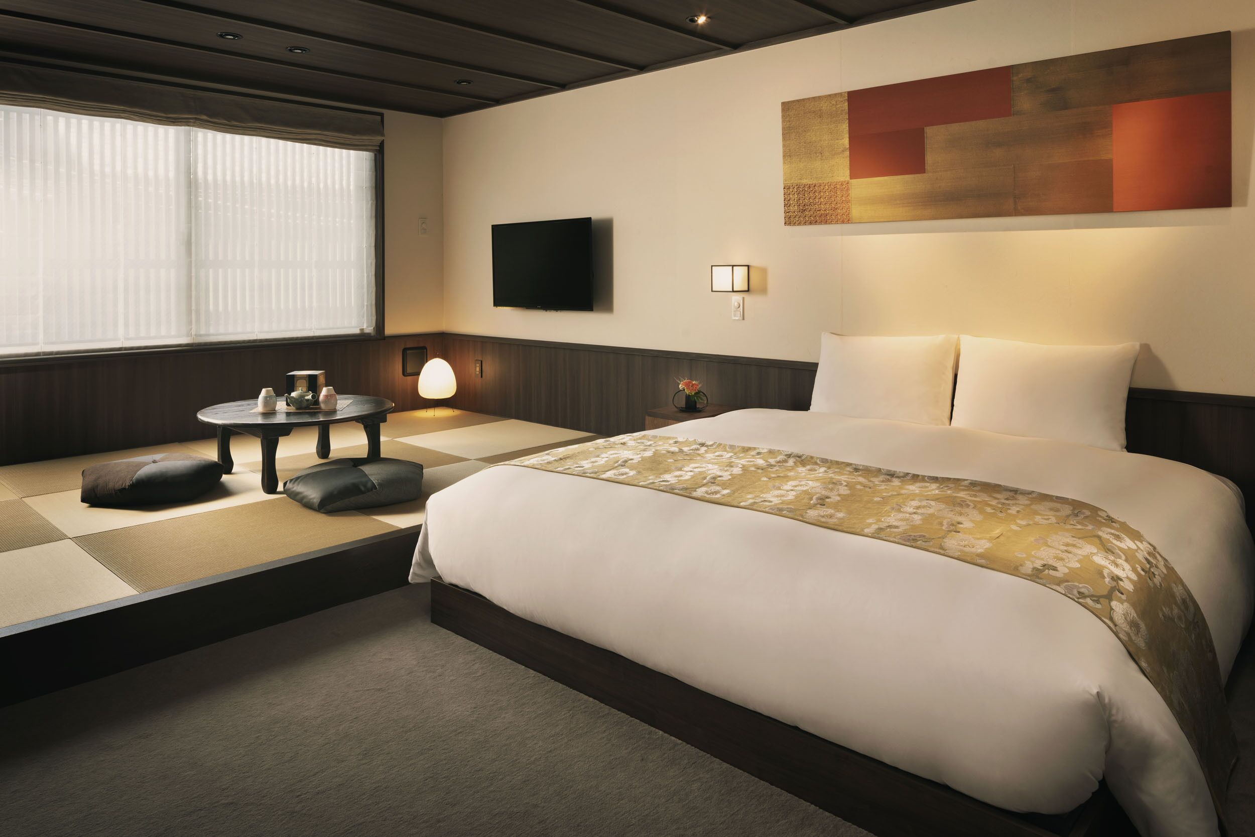 Quarto de hotel com uma cama grande, televisão, mesinha com abajur e janela com persianas.
