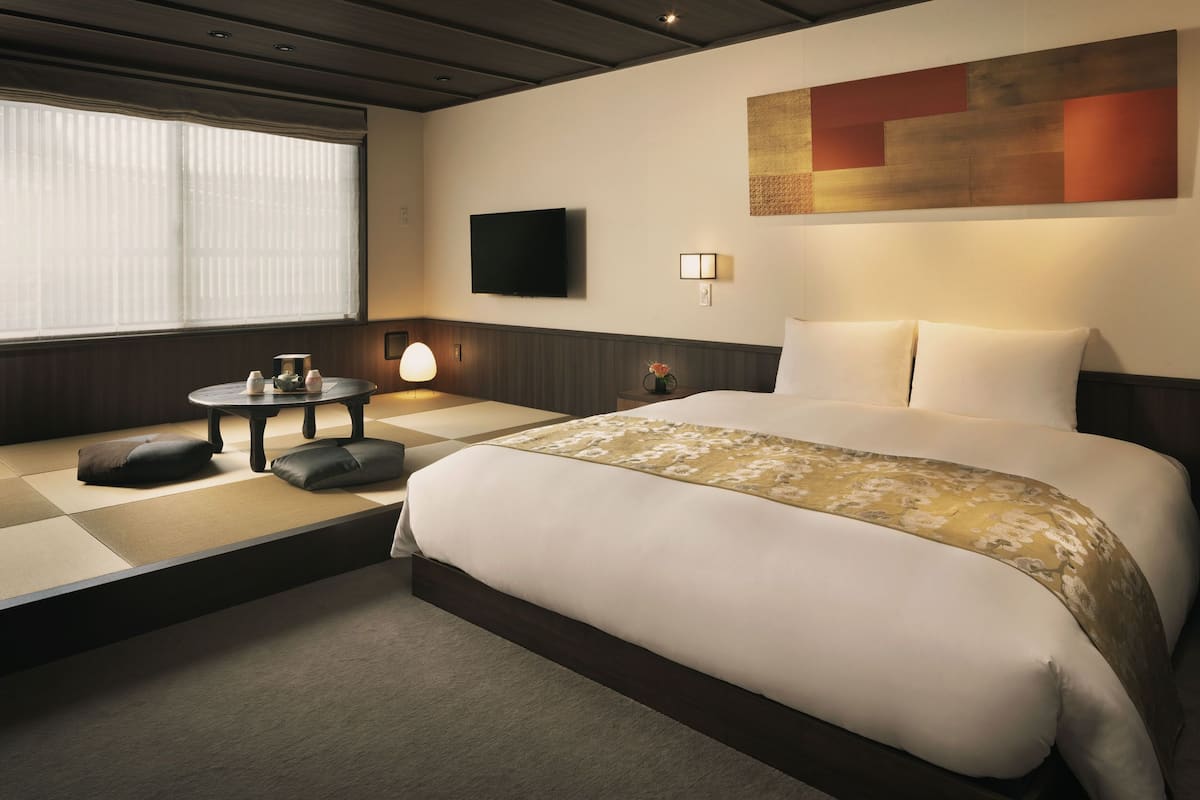 Ein Hotelzimmer mit einem großen Bett, einem Fernseher, einem kleinen Tisch mit einer Lampe und einem Fenster mit Jalousien.