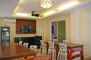 A dining area with a wooden table and white chairs, a television mounted on the wall, and a ceiling fan. - Giecel Pension (El Nido)