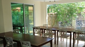 A dining area with a wooden table and white chairs, large windows with a view of greenery, and a sliding glass door leading to an outdoor patio. - Giecel Pension (El Nido)