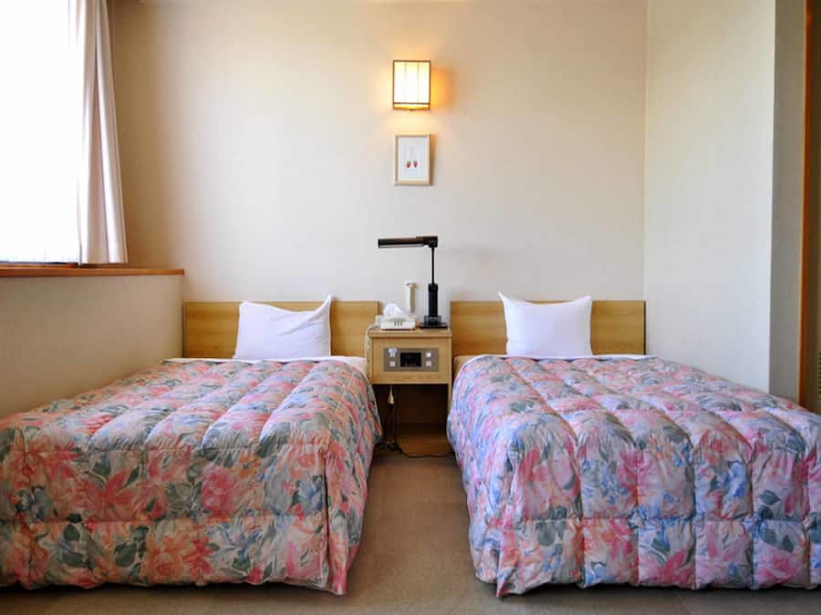 Quarto de hotel com duas camas de solteiro, cada uma com roupa de cama florida, uma mesa de cabeceira com abajur, um pequeno porta-retrato e uma mesinha de apoio.