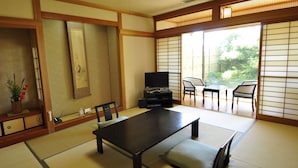 Una habitación tradicional japonesa con suelo de tatami, una mesa baja, sillas, un televisor y puertas correderas que dan a un espacio exterior.