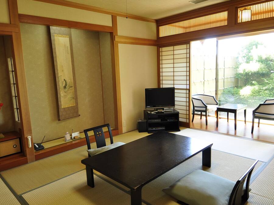 Une pièce de style japonais traditionnel, avec un sol recouvert de tatamis, une table basse, des chaises, un téléviseur et des portes coulissantes donnant sur un espace extérieur.