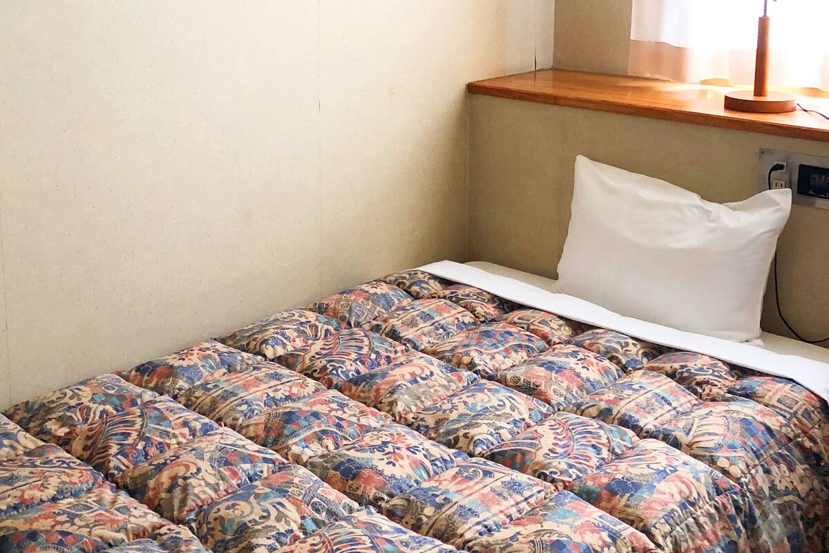 Ein Bett mit einem gemusterten Überwurf und einem weißen Kopfkissen, ein Holzregal darüber und eine Wandsteckdose.