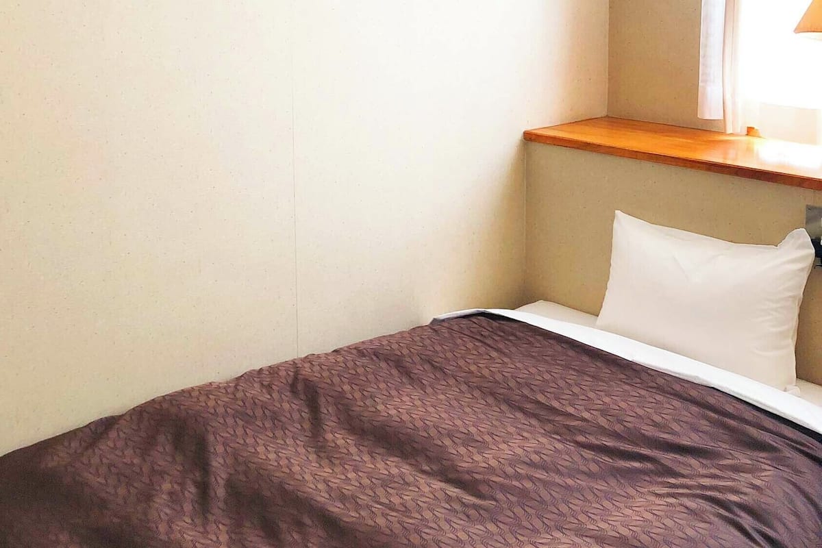 Ein Einzelbett mit brauner Decke, einem weißen Kopfkissen und einem hölzernen Kopfteil in einem kleinen, schlichten Zimmer mit hellen Wänden.