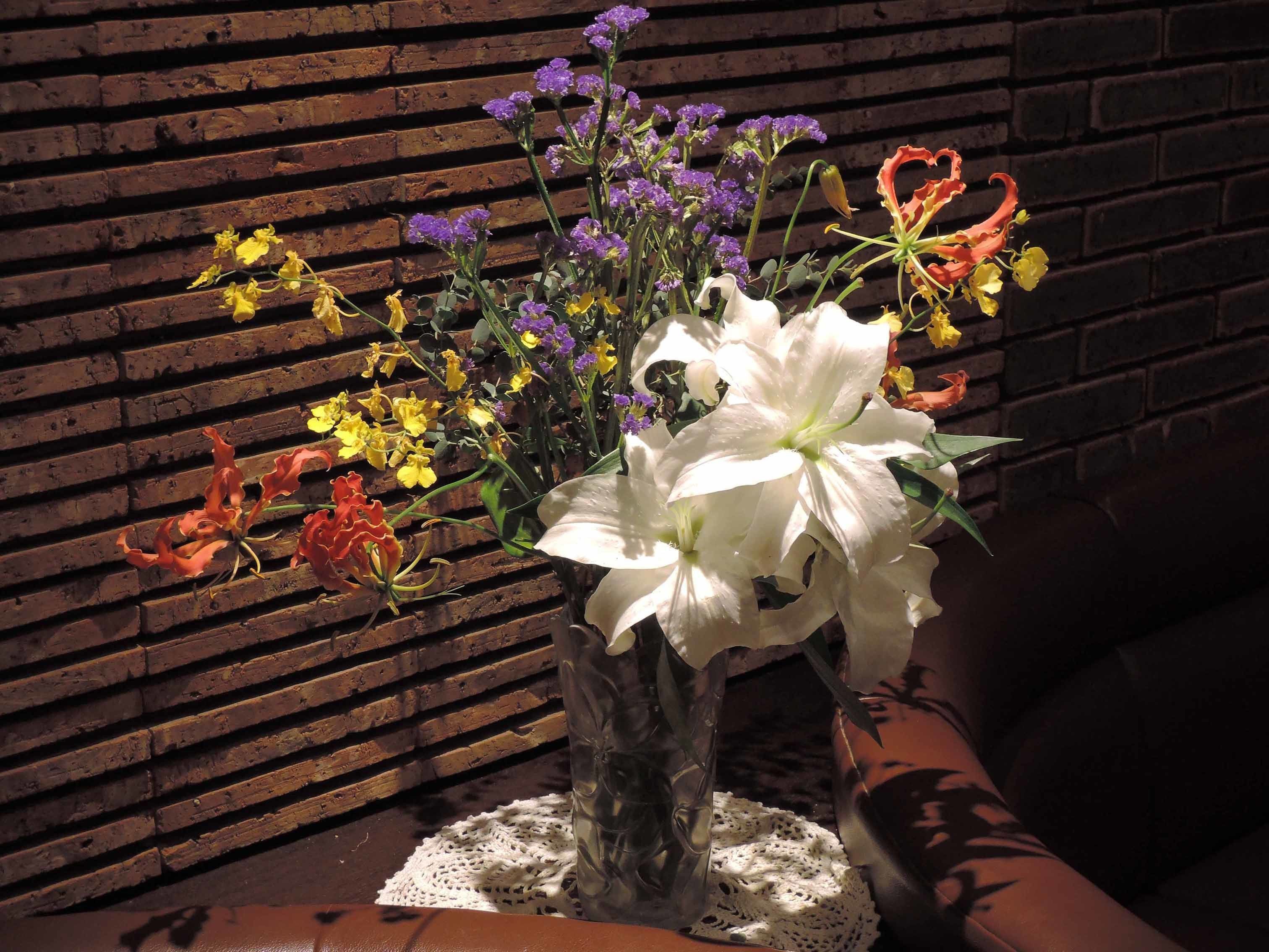 Un jarrón con diversas flores sobre una mesa, frente a una pared de ladrillo.