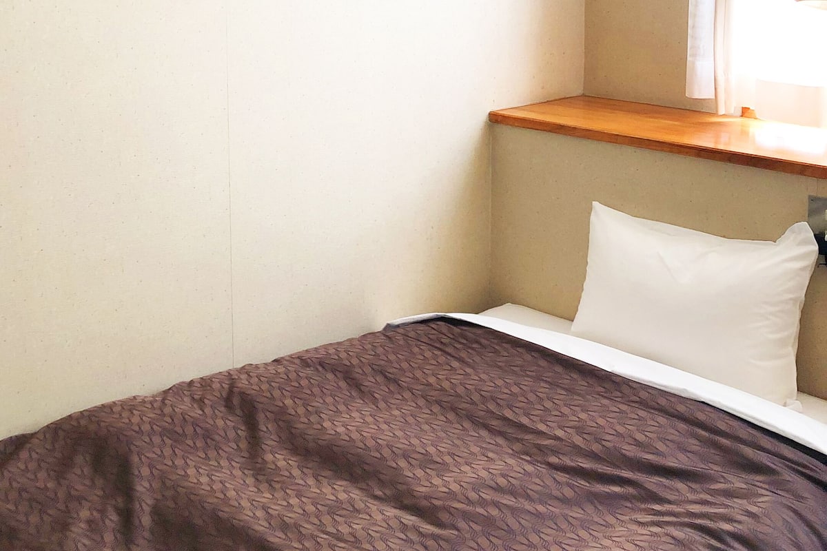 Ein kleines, ordentlich bezogenes Bett mit einer braunen, gemusterten Tagesdecke und einem weißen Kopfkissen.