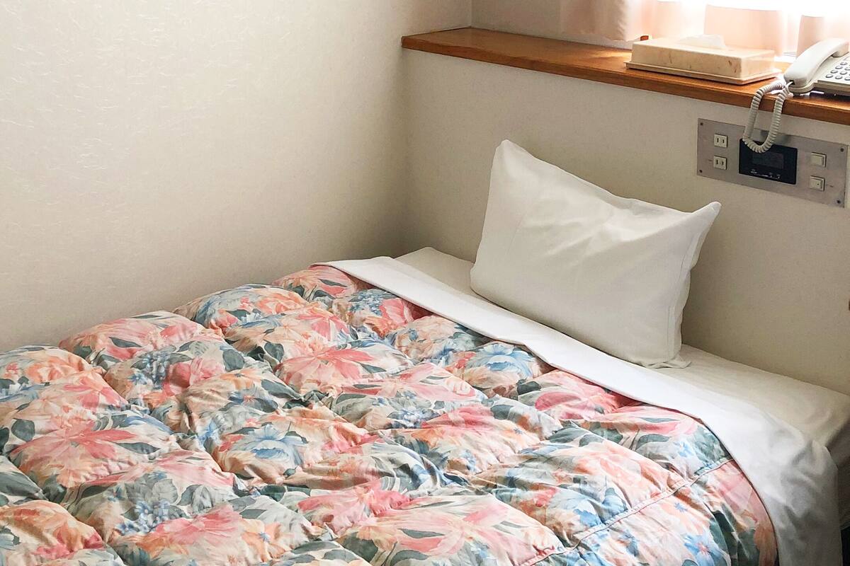 Ein Einzelbett mit einer blumigen Bettdecke und einem weißen Kopfkissen in einem Zimmer mit einem Holz-Kopfstück und einem Nachttischtelefon.