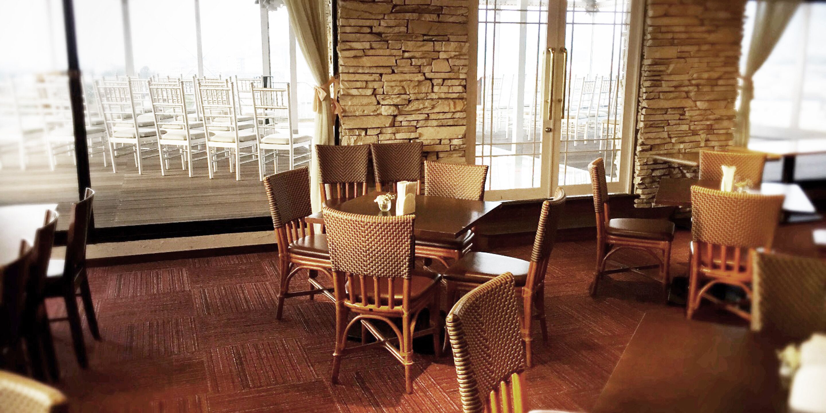 A dining area with wicker chairs and a wooden table, large windows with curtains, and a stone wall.