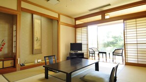 Una habitación tradicional japonesa con suelo de tatami, una mesa baja, sillas, un televisor y puertas correderas.