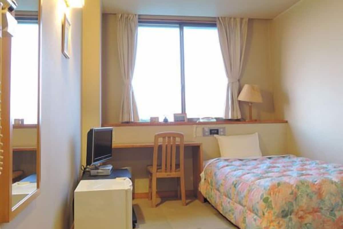 Une chambre d’hôtel comprenant un lit, un bureau avec un ordinateur, une chaise, une petite table et une fenêtre avec des rideaux.