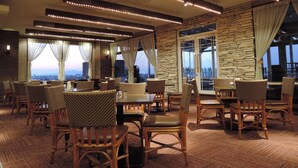 Un restaurante con sillas de mimbre, mesas, paredes de piedra y ventanas amplias con cortinas.