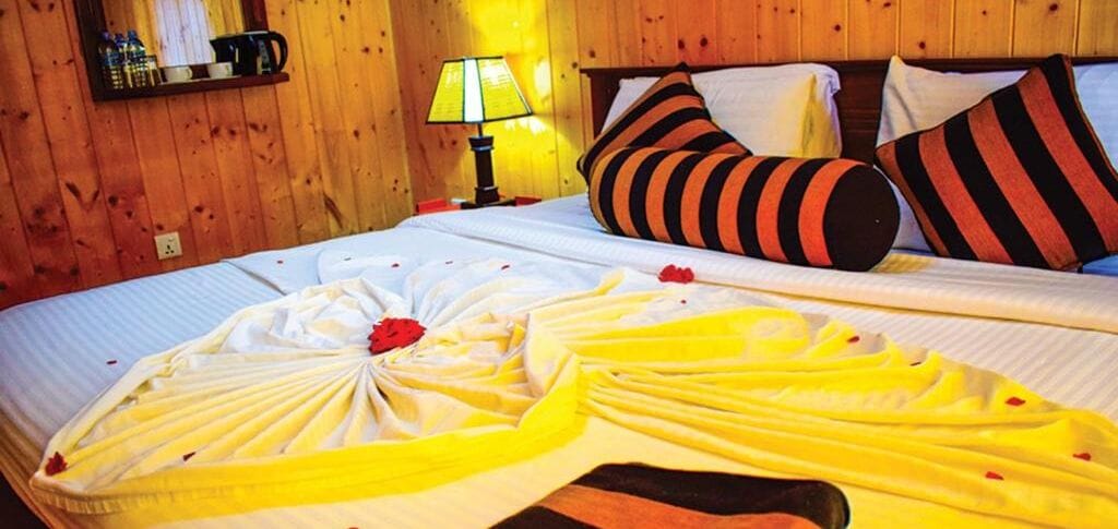 Una cama bien hecha con una toalla amarilla y una rosa roja, cabecera de madera, almohadas a rayas y una lámpara de noche.