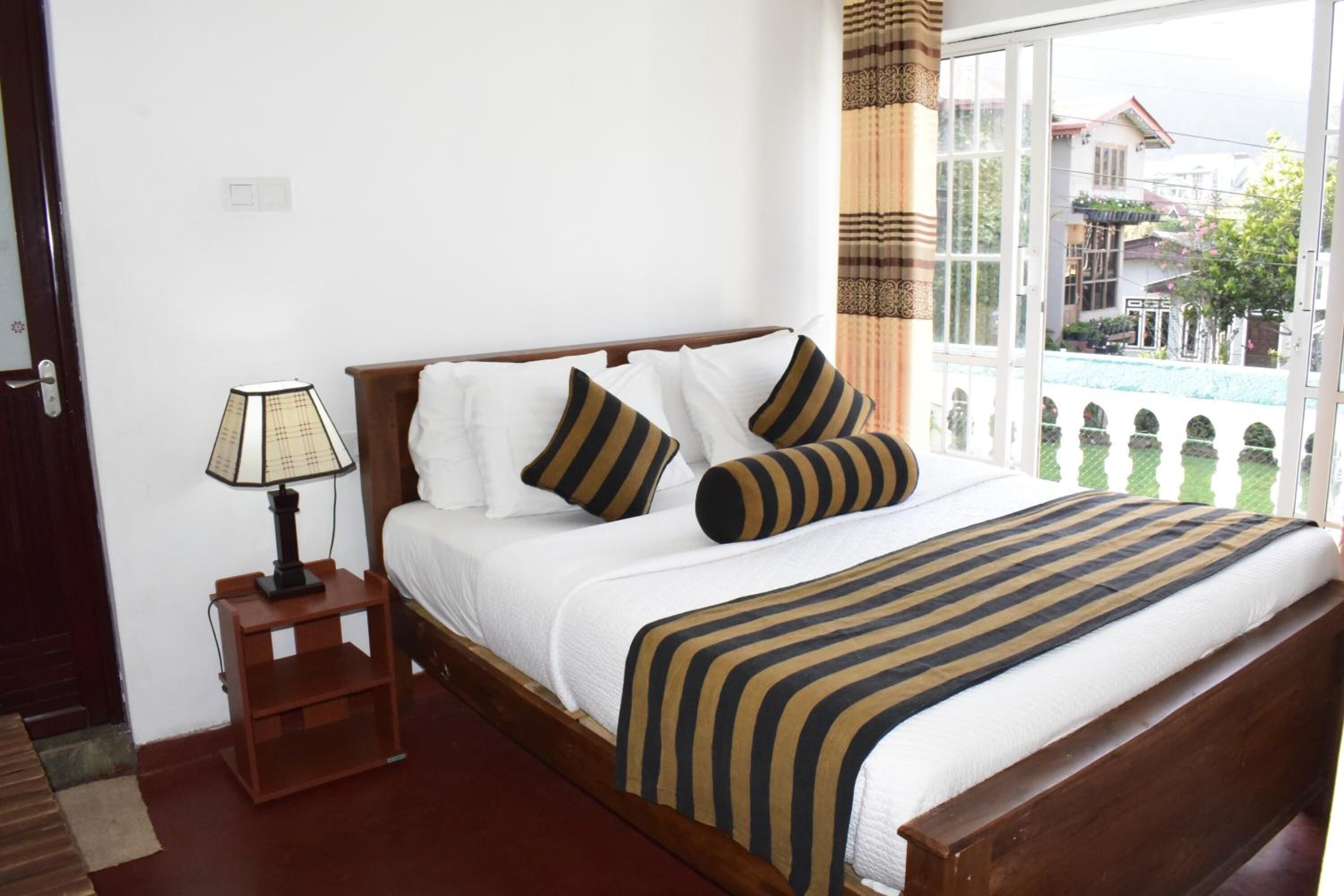A neatly made bed with white linens and striped bedding, a bedside table with a lamp, and a view of a residential area through a large window.