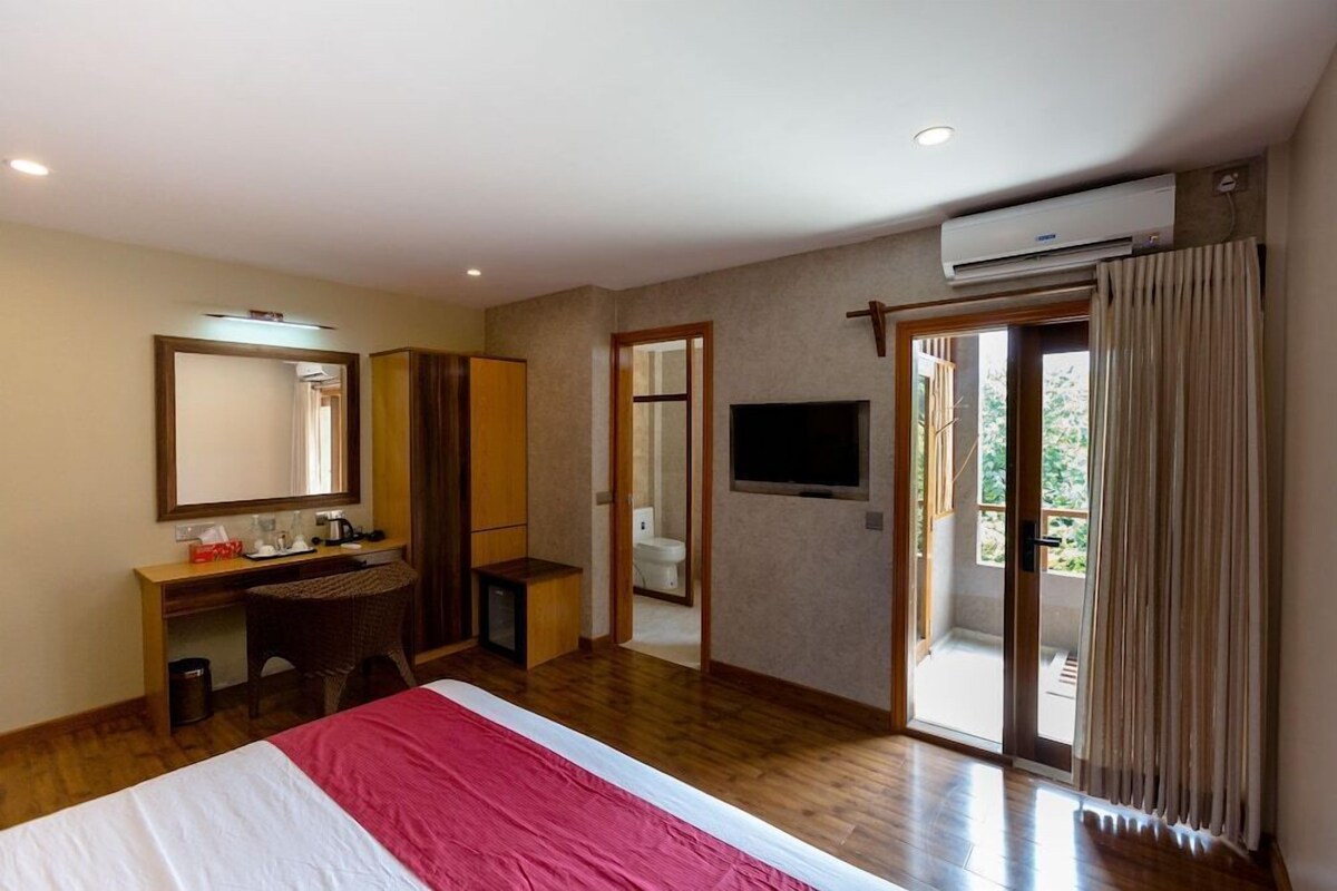 Habitación de hotel con cama, escritorio, silla, televisor, equipo de aire acondicionado y puerta corrediza a un balcón.