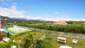 A pool area with a slide, surrounded by a white fence, and overlooking a lush landscape with mountains in the distance. - Motobu Ikoi no Yado Yamachan (Motobu)
