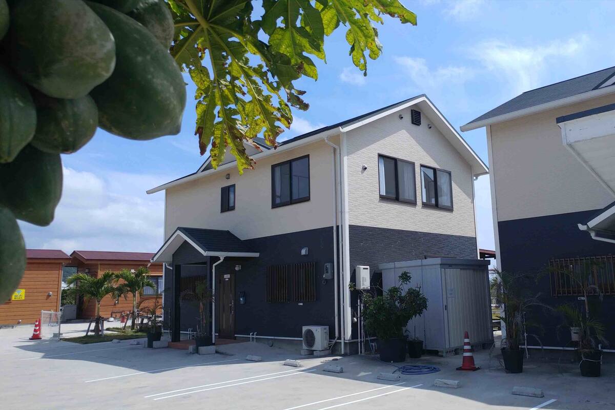 A modern two-story building with a flat roof, large windows, and a visible air conditioning unit. There is a sign with Japanese characters and a phone number on the right side.