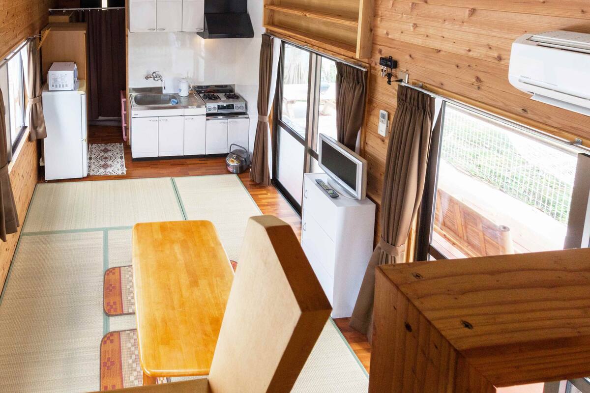 A compact kitchen with a stove, sink, and microwave. There is a dining area with a table and chairs. The room has large windows and a view of the outside.