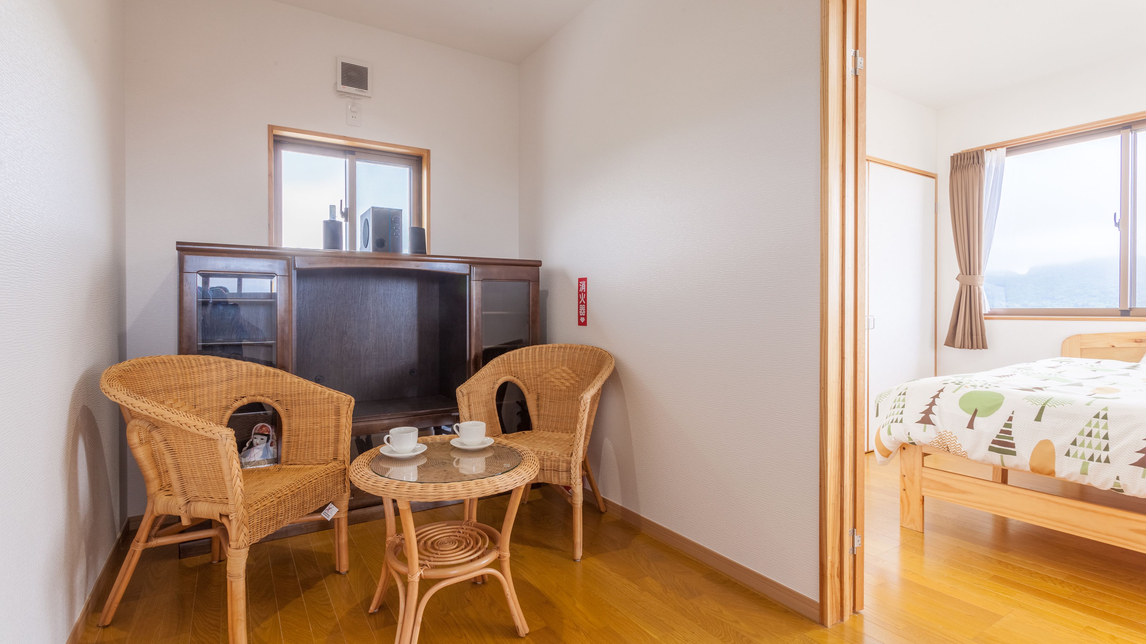 A small room with a bed, a television, two wicker chairs, a small table with cups, and a window.