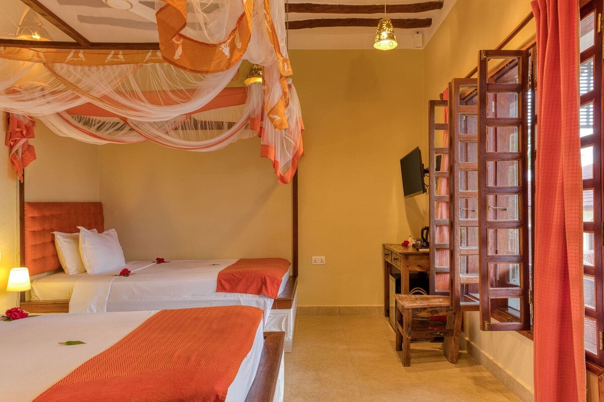 A hotel room with two beds, a wooden desk, a television, and a large window with curtains.