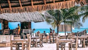 A beachside restaurant with wooden tables and chairs, a thatched roof, and a view of the ocean. - Kendwa Rocks Beach Hotel (Kendwa)