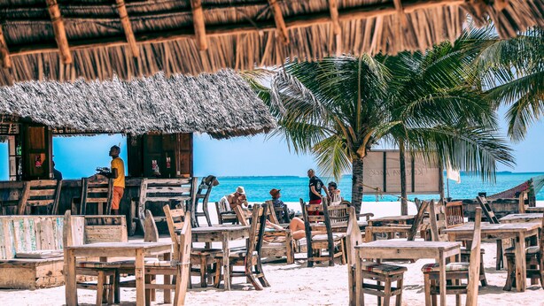 A beachside restaurant with wooden tables and chairs, a thatched roof, and a view of the ocean. - Kendwa Rocks Beach Hotel (Kendwa)