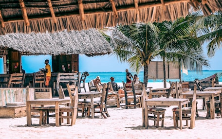 A beachside restaurant with wooden tables and chairs, a thatched roof, and a view of the ocean.