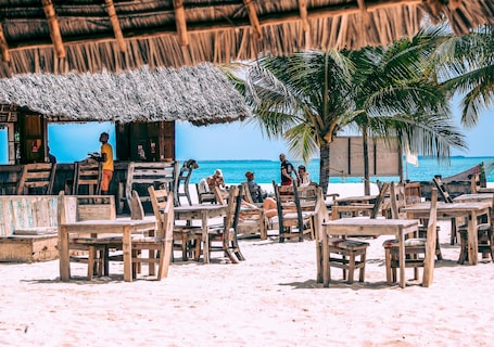 A beachside restaurant with wooden tables and chairs, a thatched roof, and a view of the ocean.