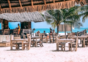 Un restaurante frente al mar con mesas y sillas de madera, techo de paja y vista al océano.