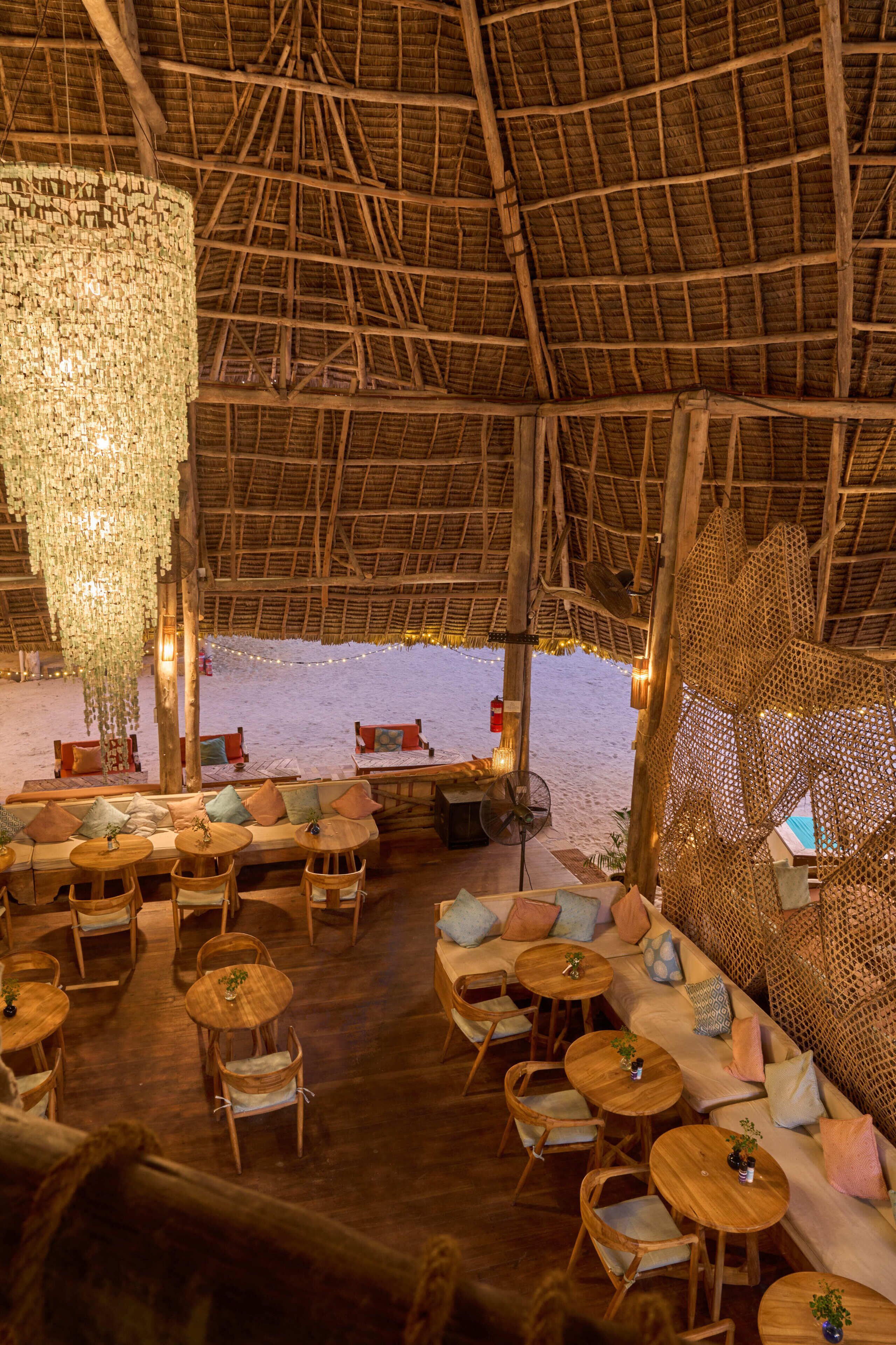 An indoor seating area with wooden furniture, a thatched roof, and a view of the beach.