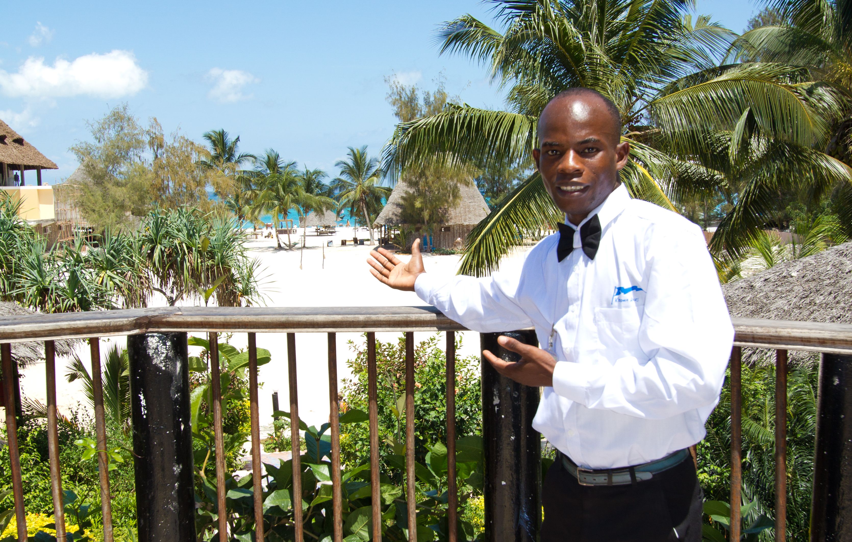 Een man in een wit overhemd en zwarte stropdas staat op een balkon met uitzicht op een strand en palmbomen.