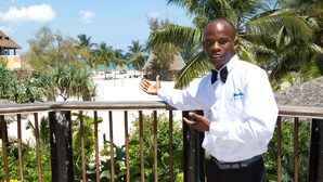 A man in a white shirt and black tie standing on a balcony with a view of a beach and palm trees. - Kendwa Rocks Beach Hotel (Kendwa)