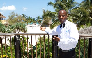 Un hombre con camisa blanca y corbata negra de pie en un balcón, con vistas a una playa y palmeras.