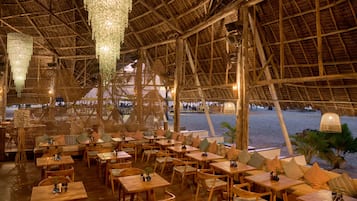 An outdoor restaurant with wooden tables and chairs, a thatched roof, and a view of the beach.