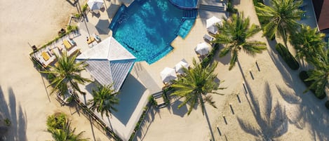 An aerial view of a resort with a swimming pool surrounded by lounge chairs and umbrellas, palm trees, and a sandy beach.