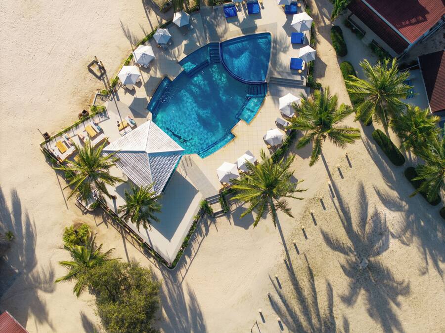 Vista aérea de un resort con piscina rodeada de sillas de descanso y sombrillas, palmeras y una playa de arena.