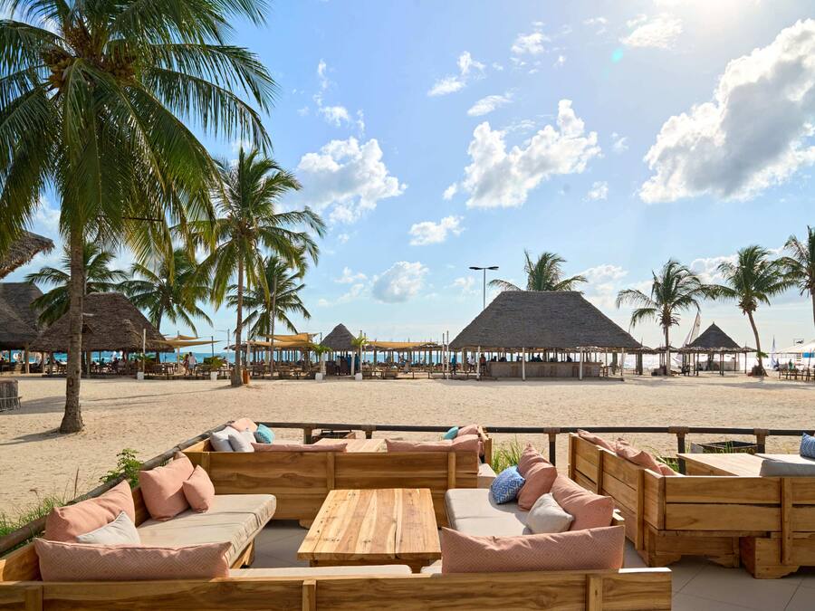 Un área de descanso en la playa con mobiliario de madera y construcciones de techo de paja.