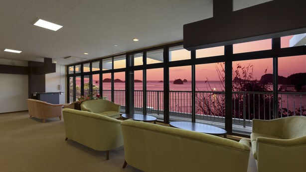 A hotel lobby with large windows offering a view of the sea and sunset. There are several armchairs and a small table. - Ikijimaso (Iki)
