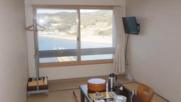 A room with a large window, a table with a kettle, a chair, and a TV on the wall.