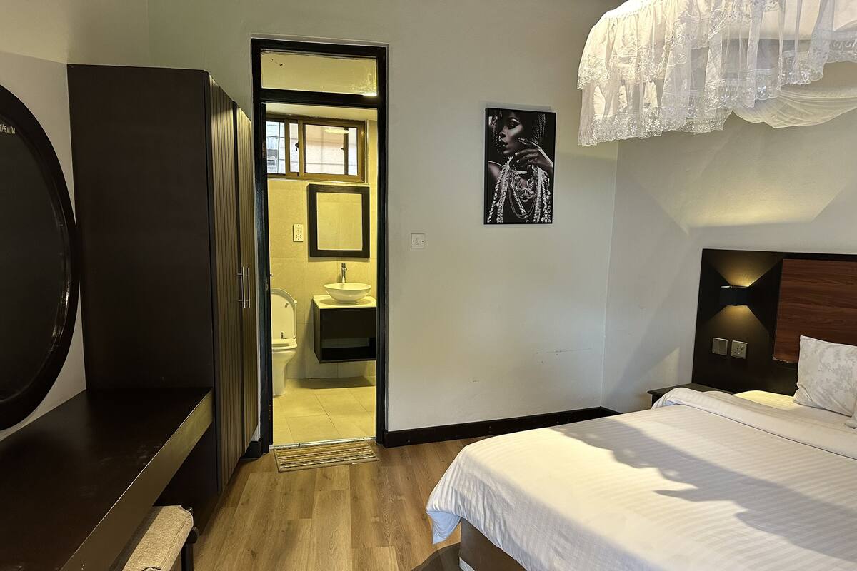 Habitación de hotel con cama, escritorio, silla, espejo y baño visible a través de una puerta abierta.