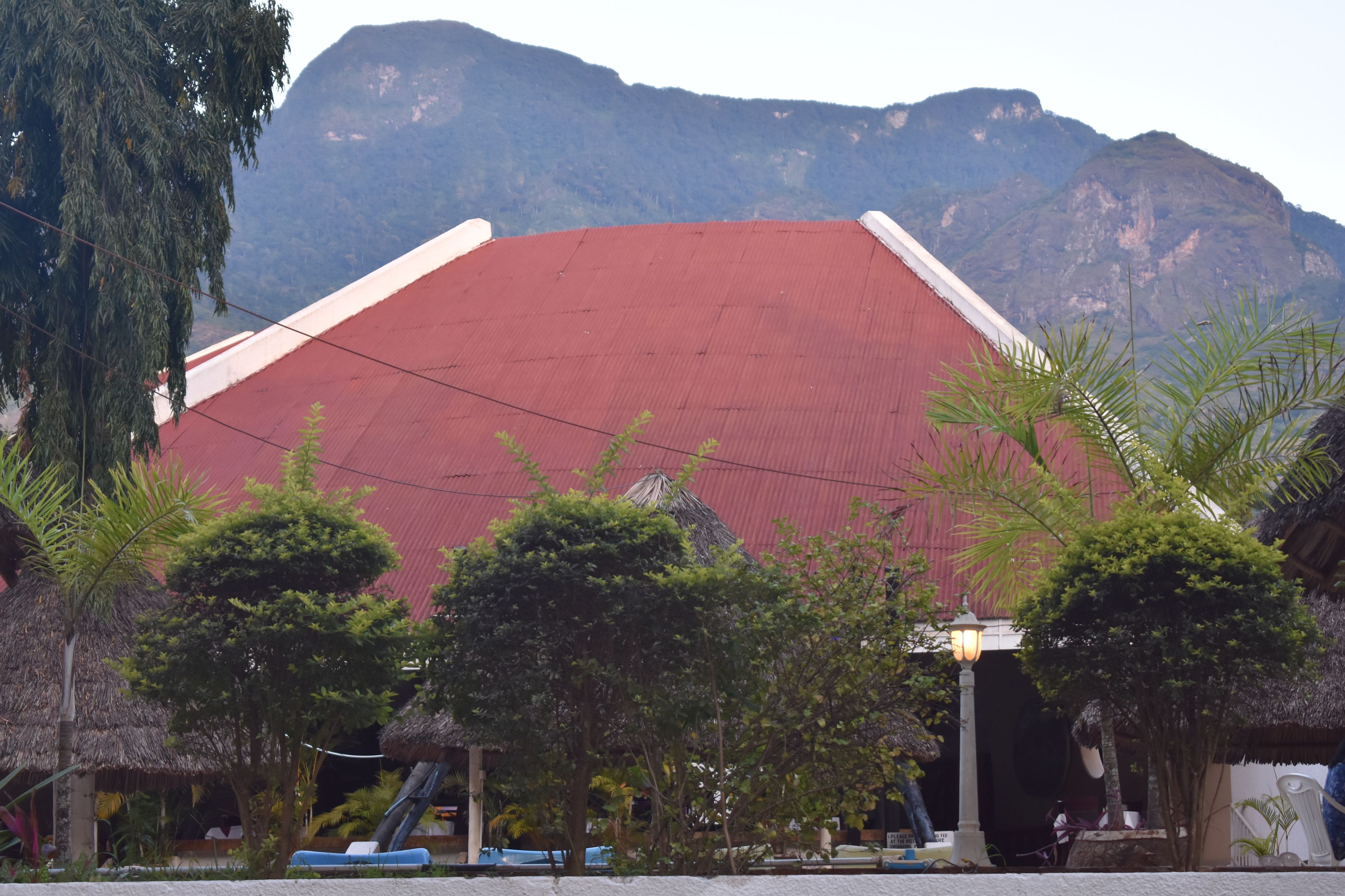 A building with a red roof and white walls, surrounded by trees and a mountain in the background.