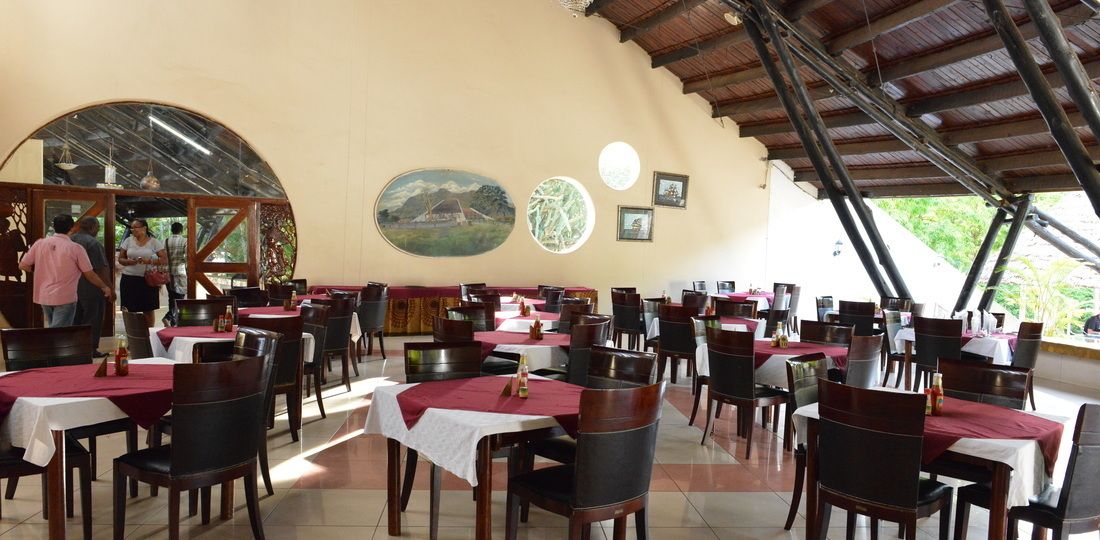 A restaurant with multiple tables and chairs, a large mural on the wall, and a view of greenery outside.