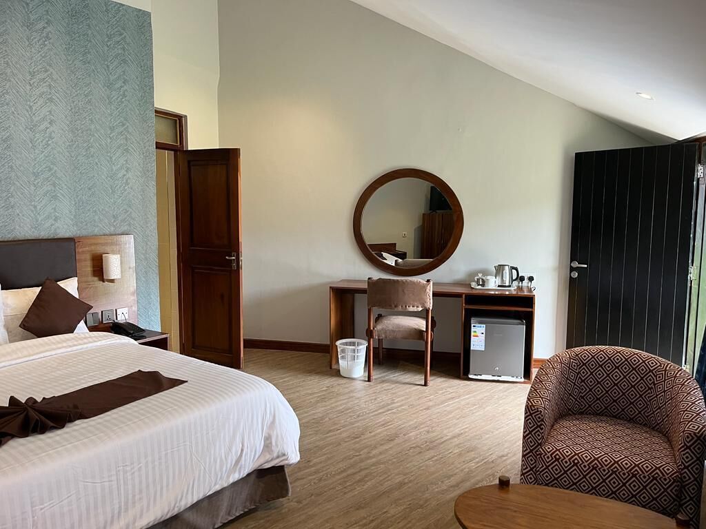 A hotel room with a bed, a desk, a chair, a round mirror, a small refrigerator, and a door.