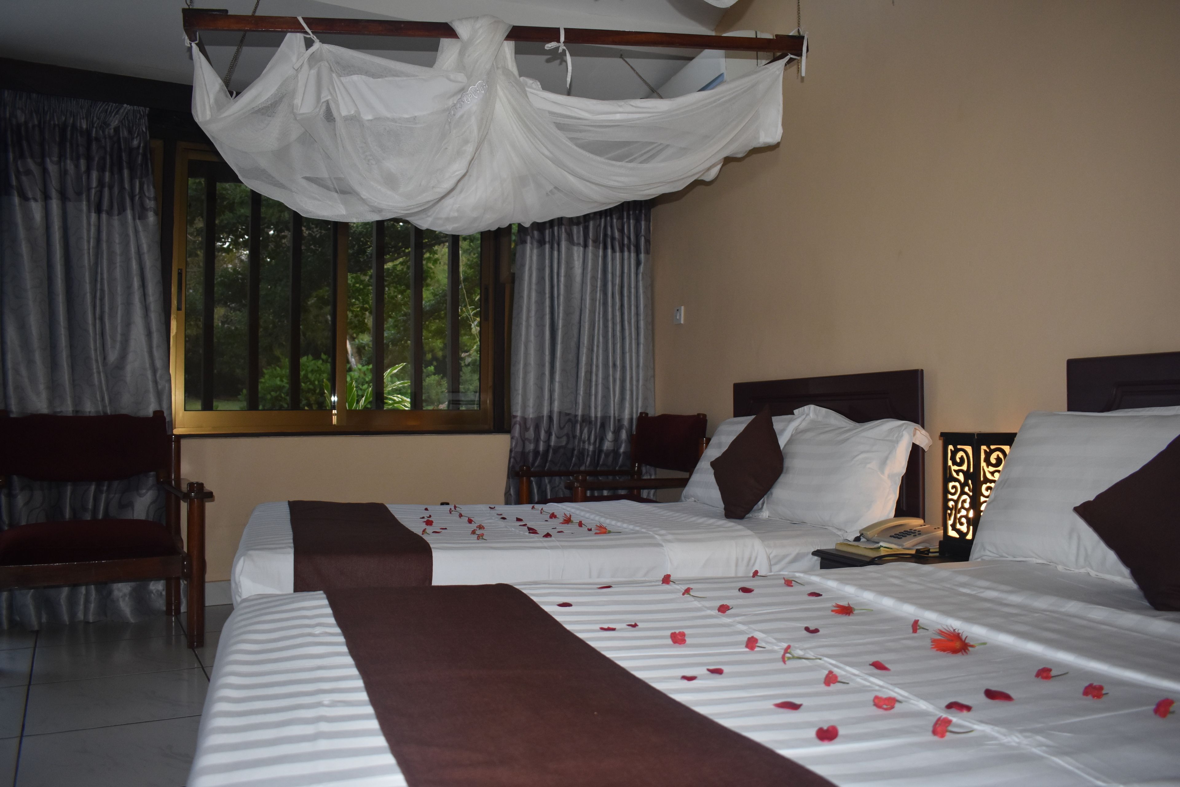 A hotel room with two beds, a canopy bed, a chair, a bedside table with a lamp, and a window with curtains.