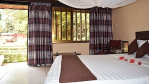 A hotel room with a large bed, curtains, a small table, and a view of greenery outside.