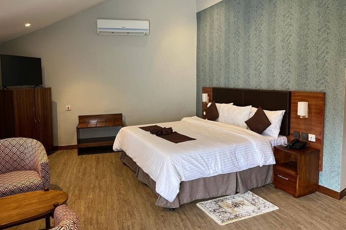 Habitación de hotel con una cama grande, cabecera de madera, mesita de noche con lámpara, un escritorio pequeño y un televisor sobre un mueble.