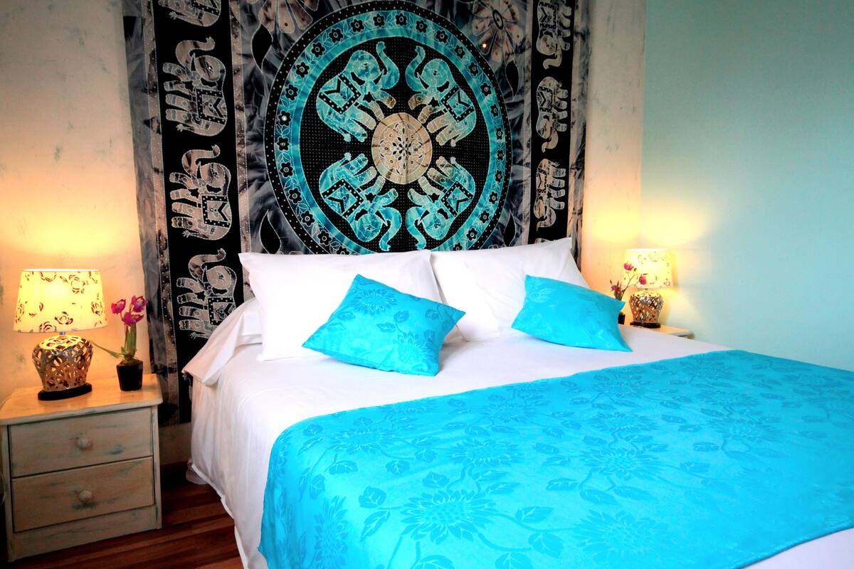 Una cama con una colcha azul estampada con motivos florales y dos almohadas a juego, un tapiz redondo con un diseño circular central, y dos lámparas de noche con patrones florales.