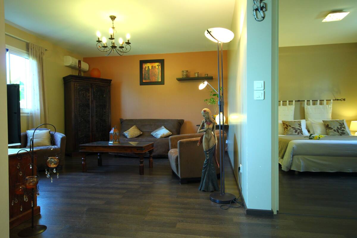 Una sala de estar con un sofá, un sillón y una mesa. Se observa una estatua, una lámpara y una ventana con cortinas.