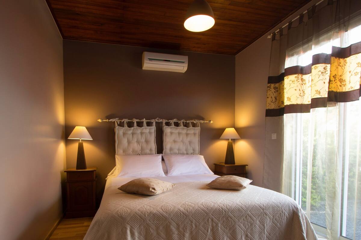 Un dormitorio con una cama, dos mesitas de noche con lámparas, una lámpara de techo, un equipo de aire acondicionado y una ventana con cortinas.