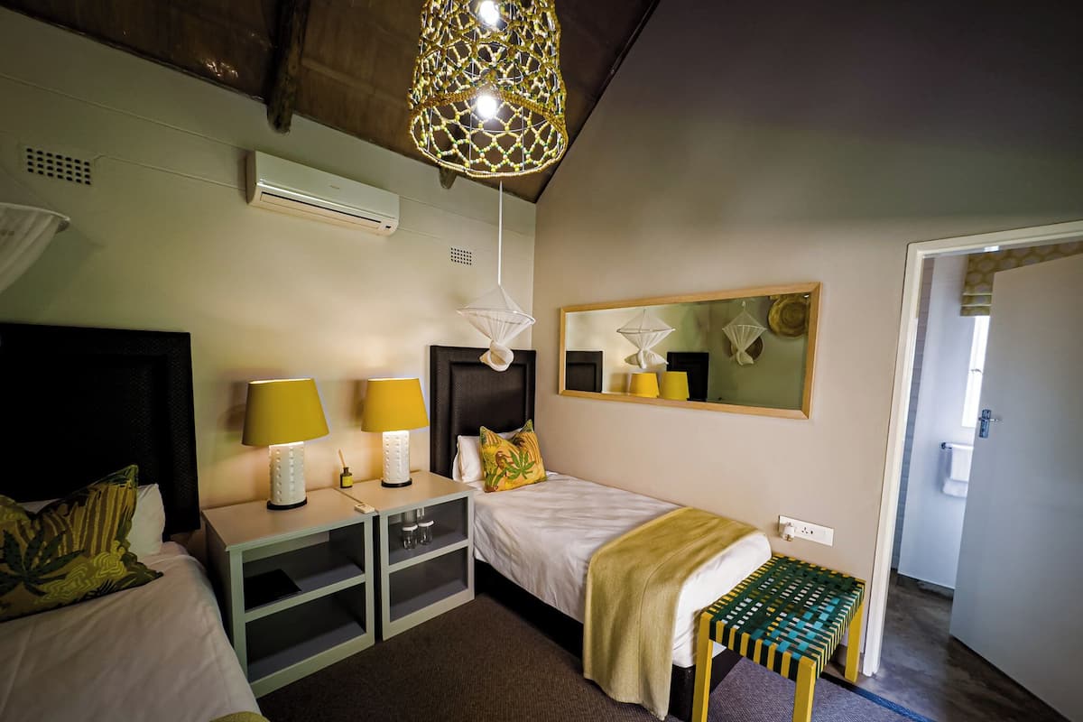 Quarto de hotel com cama, abajures, uma pequena mesa, um banquinho, espelho e ar condicionado.